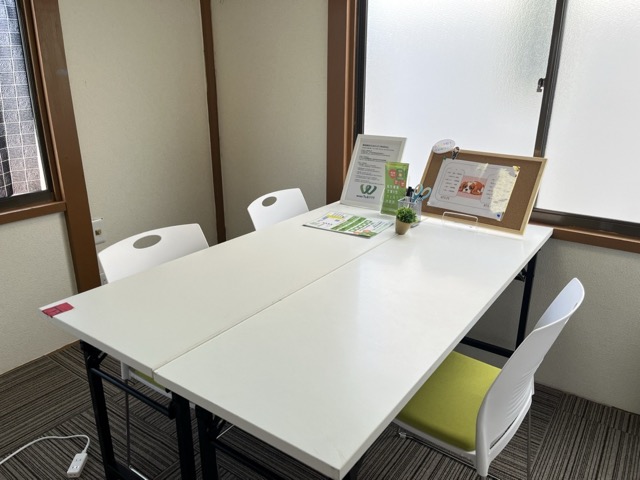 窓際に白いテーブルと椅子が並んだ会議スペース。案内資料や掲示物が置かれている